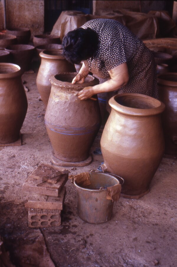 At the Kornos Pottery Cooperative workspace, Eleni Alecou walked around an oven to form a rim from the last coil. Blue yarn encircling the lower body constrict the wet clay. Nearly finished ovens without yarn had a small spout-like flue. June 1986. Photo by the Author.