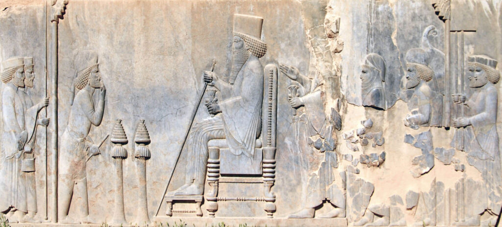 Fig. 4 The Audience Scene on the Persepolis Treasury: The Great King (Darius I or Xerxes I) is sitting on the throne while holding a long scepter in his right hand. (Wikimedia Commons / Pawel Ryszawa)