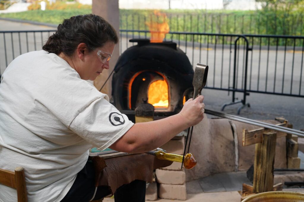 Corning Museum Hot Glass Demonstrator, Catherine Ayers, shapes glass using jacks and a thigh-board at the reproduction ancient wood-fired glass furnace. Image Courtesy of The Corning Museum of Glass.