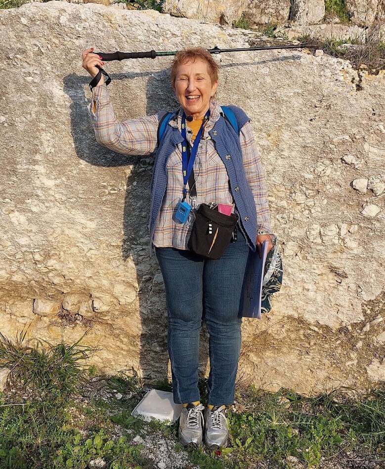 Norma Franklin demonstrating the height of the 4m-high artificial scarp separating the royal palace at Sebaste from the rest of the surrounding area. She is 1:50 meters tall so ca 2 . 5 meters of the rocky scarp is now buried by earthen debris washed since the area was excavated in 1910. (Photo by M. van den Berg)