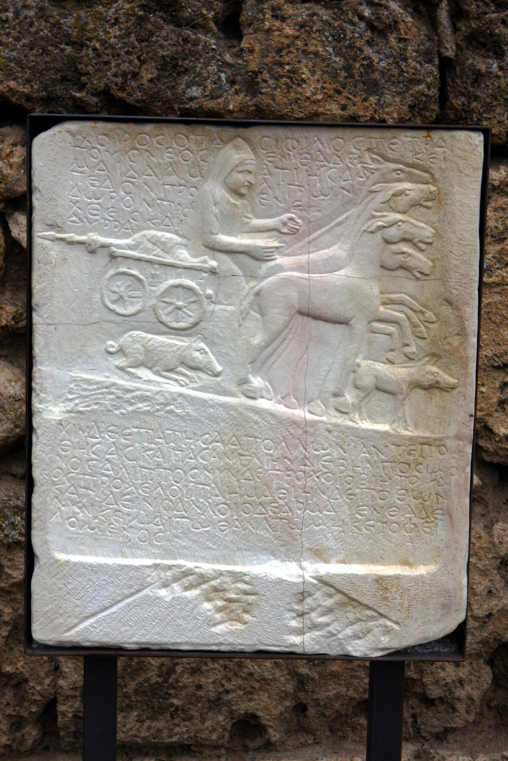 Copy of the “pig stele” of Edessa with Greek inscription (original in the Edessa Archaeological Museum). The accompanying relief shows the pig twice: on the right, it is walking along before the accident, while on the left it is lying on the ground after being crushed by the wheels of the chariot, its skin ruffled in mortal agony. Photo by Philipp Pilholfer. CC-BY-SA 3.0.