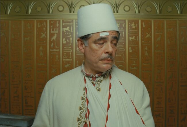 Korda (Benicio del Toro), wearing a white fez and a long white garment ormanented with stylized papyrus fronds. Also note the Egyptian hieroglyphic decoration of the elevator background. Still image from The Phoenician Scheme (2025), directed by Wes Anderson. Courtesy of Focus Features.