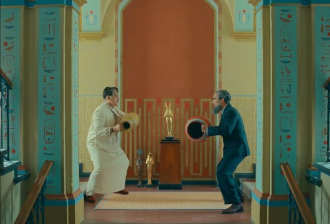 Korda (in white robe) and Uncle Nubar (in a dark pinstripe suit) fight in the stairwell of the “Royal Majestic Imperial Calouste Korda Desert Oasis Palace.” Still image from The Phoenician Scheme (2025), directed by Wes Anderson. Courtesy of Focus Features.